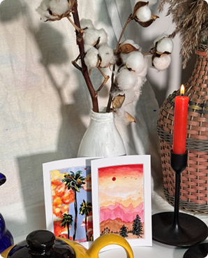 Two palm-tree sunset postcards propped near dried cotton stems and a red candle
