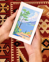 Vibrant postcard of a Mediterranean coastline and blue bay held against a kilim rug