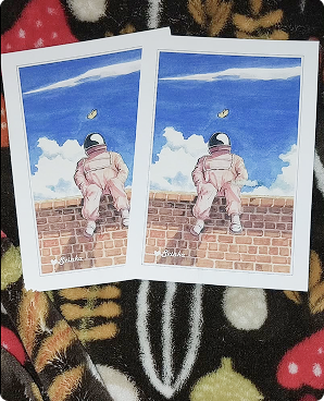 Two astronaut-on-a-brick-wall postcards lying on dark floral fabric